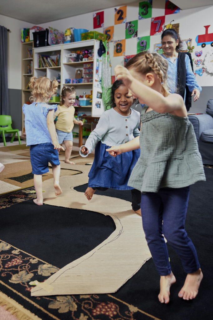 Group of four young girls laughing and playing in a playroom, with an adult woman smiling and supervising close by