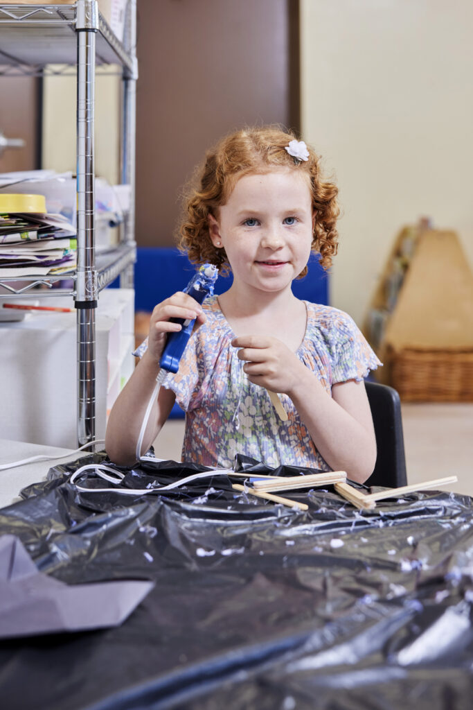 Small girl playing with a glue gun and popsicle sticks