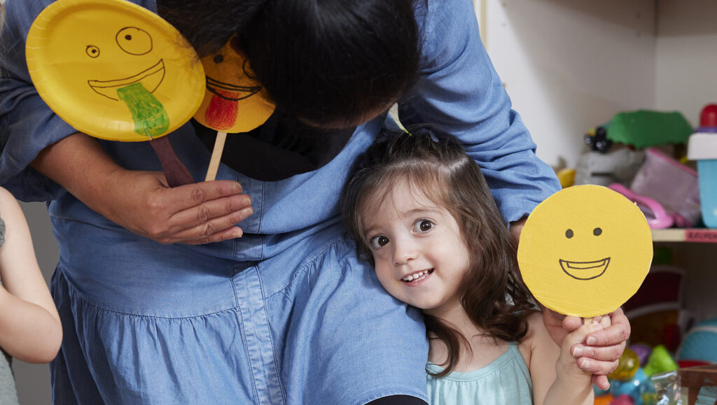 Young girl hugging teach and holding up paper smiley faces
